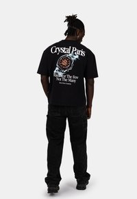 Černé bavlněné tričko s grafickým designem na zadní straně, které obsahuje text a obvodový vzor. Kombinováno s černými cargo kalhotami a botami.