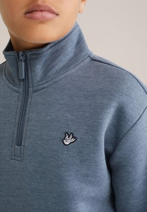 Plavi pulover na zip s visokim ovratnikom, napravljen od mekanog materijala. Ima izvezeni logotip ptice na prsima. Glatka tekstura.