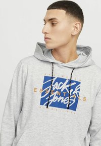 Sudadera gris con un gran diseño gráfico en azul y naranja que presenta 'Jack & Jones' y cordones. Tejido suave con un bolsillo tipo canguro.