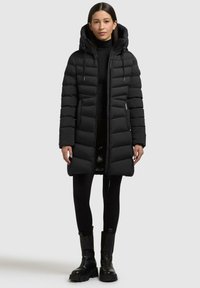 Piumino nero con cappuccio, caratterizzato da una texture trapuntata, zip frontale, stringhe regolabili e tasche laterali. Abbinato a leggings e stivali.