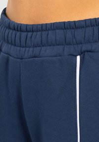 Marineblaue Sweatpants mit einem strukturierten, elastischen Bund, Seitentaschen und kontrastierendem weißen Piping entlang der Nähte.