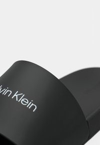 Infradito nere con una fascia larga e il logo bianco "Calvin Klein" impresso. Materiale liscio e suola piatta. Design minimalista, senza motivi.