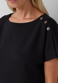 Zwarte top met korte mouwen en een ronde halslijn, voorzien van drie metallic drukknoopjes op de linker schouder en een zachte, gladde textuur.