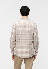 Άνδρας που φοράει μπεζ και λευκό πουκάμισο με καρό μακριά μανίκια και χακί παντελόνι, γυρισμένος πλάτη σε απλό ανοιχτό φόντο.