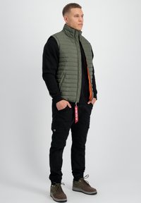 Olivgrön kviltad väst med hög krage, orange foder, i kombination med svarta cargobyxor och bruna sneakers. Har dragkedja och etiketter.