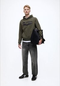 Olivově zelená mikina s kapucí s potiskem loga, šedé džíny a černé tenisky. Model drží černý batoh a stojí před bílým pozadím.