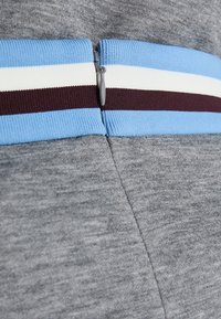 Un tissu gris à la texture lisse présente une bande élastique rayée bleue, blanche et bordeaux ainsi qu'une fermeture éclair grise à la taille.
