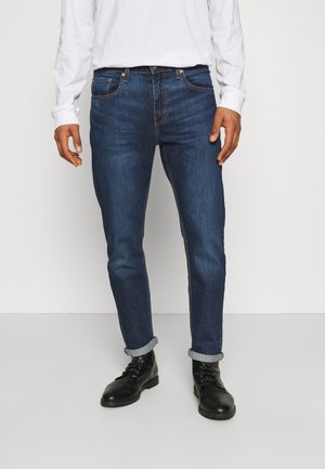 Homme portant un jean slim bleu foncé avec revers, une chemise blanche à manches longues et des bottes noires à lacets, debout devant un fond clair uni.