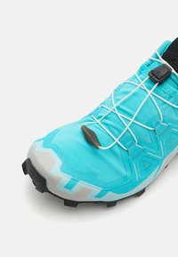 Turkos färgad trail-löparskor med syntetiskt ovandel, vita elastiska skosnören och en strukturerad grå gummisula. Utrustad med ett säkert dragstängningssystem.