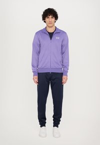 Violets džemperis ar rāvējslēdzēju un augstu apkakli, pieguļošiem piedurkņu galiem un logotipu; kombinēts ar tumši ziliem biksēm un baltiem sneakers. Gluda auduma, ikdienas piegriezuma.