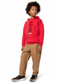 Bambino con capelli ricci che indossa un felpa rossa con il testo "Sii il tuo capo", pantaloni beige e sneakers nere e marroni, in piedi di lato.