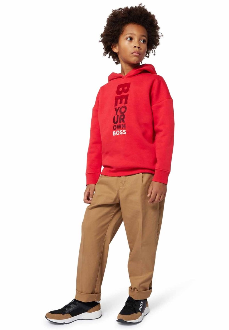 Bambino con capelli ricci che indossa un felpa rossa con il testo "Sii il tuo capo", pantaloni beige e sneakers nere e marroni, in piedi di lato.