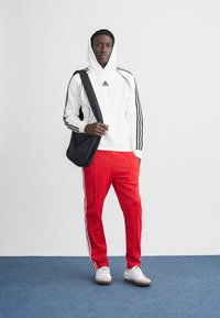 Bijela majica s kapuljačom s crnim prugama i logotipom, crvene trenirka hlače sa bočnim prugama i bijele tenisice. Crna torba prebačena preko ramena.