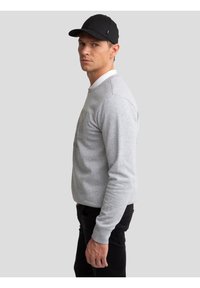 Mand iført sort kasket, lysegrå sweatshirt, hvid skjortekrave og sorte bukser, stående sideværts mod en ensfarvet grå baggrund.