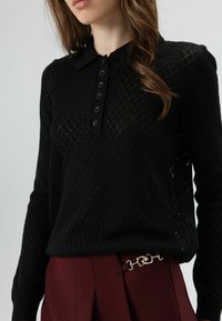 Zwart gebreide trui met diamantpatroon, kraag met knoopsluiting aan de voorkant. Gecombineerd met bordeauxrode broek met zilveren kettingdetail.