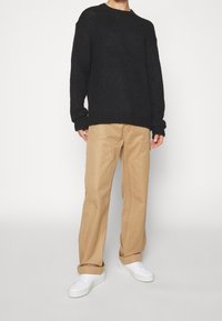 Czarny sweter dzianinowy z prążkowanymi mankietami i dołem, beżowe spodnie z widocznymi szwami oraz białe trampki, prezentowane na jednolitym tle.