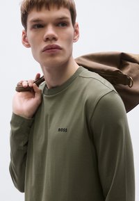 Μακρυμάνικο πουκάμισο σε ελιά πράσινο με στρογγυλή λαιμόκοψη και μικρό μαύρο λογότυπο "BOSS" στο στήθος. Το μοντέλο κρατά καφέ τσάντα στο ύψος του ώμου.