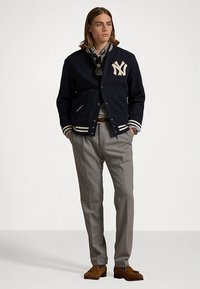 Námořnicky modrá vlněná univerzitní bunda s krémovými pruhy a logem NY, nošená přes světlou košili; doplněná šedými kalhotami a hnědými mokasínami.