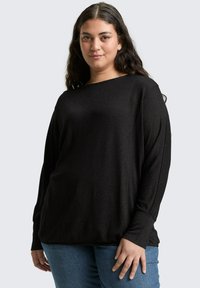 Zwarte lange mouwen shirt met een losse pasvorm, bootnek en geribde manchetten, gecombineerd met blauwe jeans. Zachte textuur met een subtiele glans.