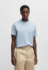 Hellblaues, strukturiertes T-Shirt mit Rundhalsausschnitt und kurzen Ärmeln, kombiniert mit beigen Hosen, die einen Knopfdetail aufweisen.