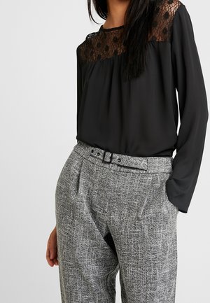 Femme portant un chemisier noir à manches longues avec empiècement en dentelle et un pantalon gris texturé taille haute avec ceinture assortie, main dans la poche.