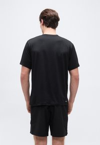 Άνδρας που στέκεται με την πλάτη, φορώ ντας απλό μαύρο κοντομάνικο μπλουζάκι και μαύρο σορτς, μπροστά σε απλό ανοιχτόχρωμο φόντο.