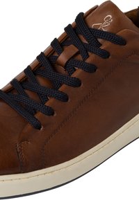 Adidași din piele maro cu sireturi negre, având o talpă din cauciuc bej care contrastează și detalii subtile de cusătură.