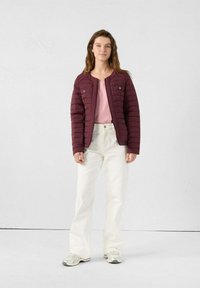 Bordeaux gewatteerde jas met een horizontaal gestikt patroon, voorzien van drukknoopsluitingen en twee voorzakken, gecombineerd met een roze top en witte broek.