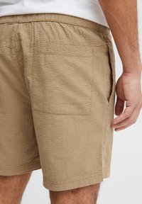 Beige katoenen korte broek met een gestructureerd oppervlak, een elastische tailleband, steekzakken aan de zijkant en een achterzak. Afgewerkte randen zonder extra versieringen.