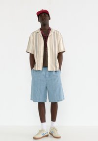 Chemise à manches courtes rayée en crème et gris, associée à un short à fines rayures bleu clair. Les accessoires comprennent un bandana bordeaux et une casquette.