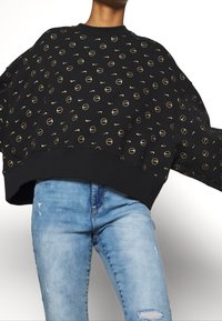 Black sweatshirt with gold circular and Nike logo patterns, oversized fit, ribbed hem, paired with light blue distressed jeans.