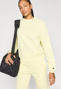 Κίτρινο φούτερ με ριμπ μανσέτες και τελείωμα, με μικρό λογότυπο. Συνδυασμένο με παντελόνι στο ίδιο χρώμα, κρατώντας μια μαύρη τσάντα με υφή και λουράκι.