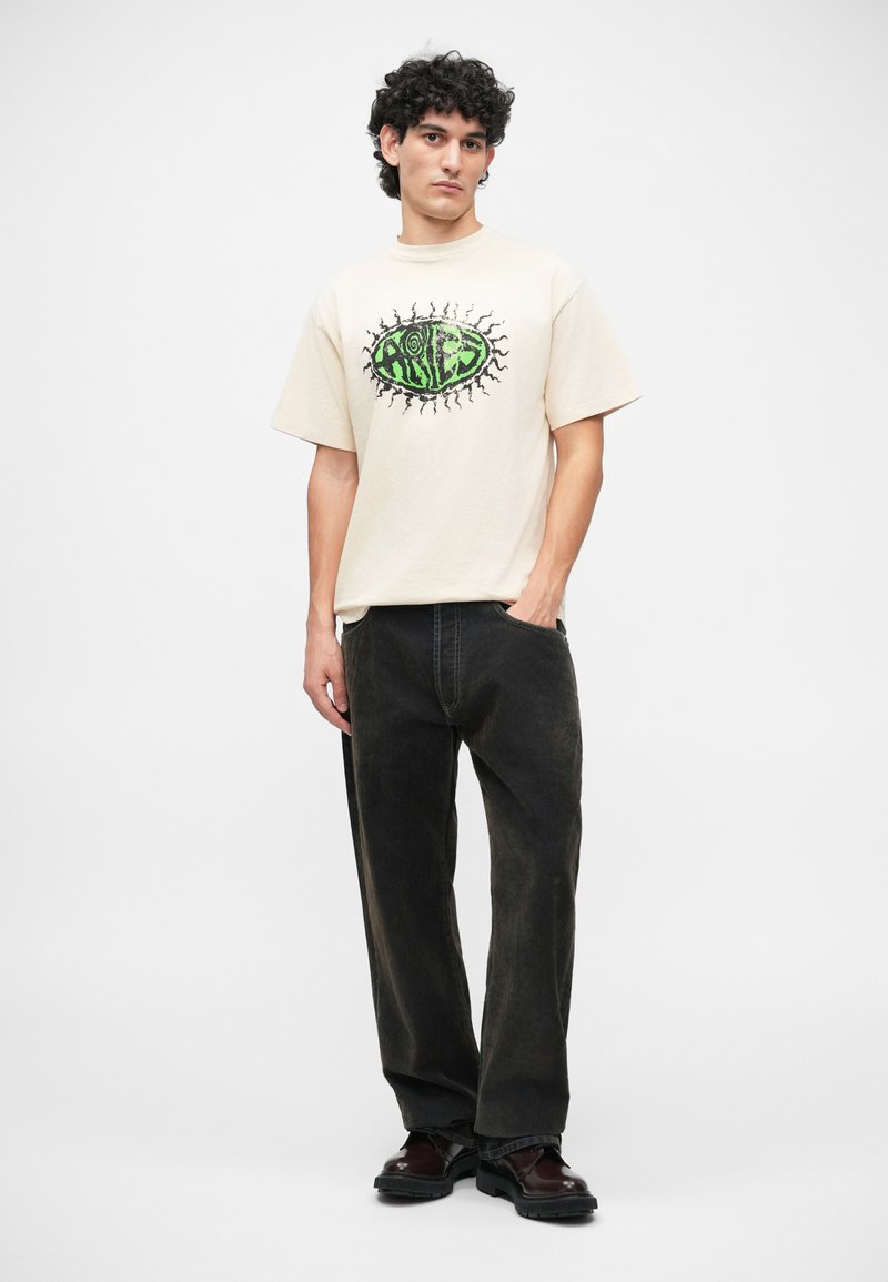 Tricou bej cu mânecă scurtă, având un design grafic verde și negru, asortat cu blugi largi de culoare închisă și pantofi negri.