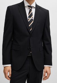 Completo scuro in navy con vestibilità su misura, dotato di chiusura a due bottoni, rever a punta e tessuto leggermente texture. Abbinato a una cravatta a righe.