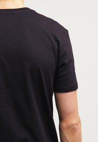 Persona vista desde atrás, llevando una camiseta negra lisa de manga corta sobre un fondo claro y sencillo.