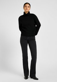 Frau in einem schwarzen Rollkragenpullover, schwarzen geraden Jeans und schwarzen spitzen Stiefeln, die vor einem schlichten Hintergrund steht.