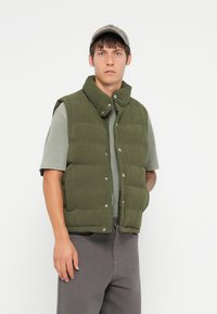 Vesta puffer verde din catifea cu guler înalt și nasturi, purtată peste un tricou gri, asortată cu pantaloni gri. Design minimalist.