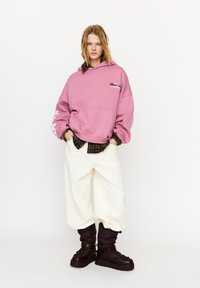 Felpa oversize rosa con logo sul petto, indossata sopra una camicia scura a quadri, abbinata a pantaloni larghi color avorio e stivali marroni robusti.