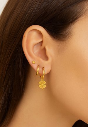 Goldene Creolen mit einem Blattanhänger; glänzende Textur, mehrschichtiges Design, kleines Ohrring-Stift daneben; vor einem Haut-Hintergrund platziert.