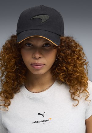 Młoda kobieta o kręconych włosach, ubrana w czarną czapkę McLaren oraz białą koszulkę Puma McLaren Racing na neutralnym szarym tle.