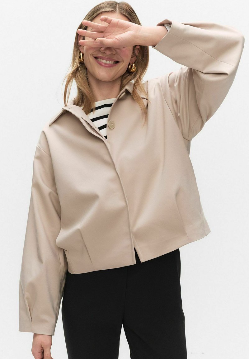 Woman smiling with hand partially covering her face, wearing beige jacket, black pants, striped shirt, and gold hoop earrings.