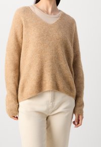 Světle hnědý svetr s výstřihem do V vyrobený z měkké texturované tkaniny. Má dlouhé rukávy, uvolněný střih a mírně zkrácený lem. Je kombinován se světlými béžovými kalhotami.