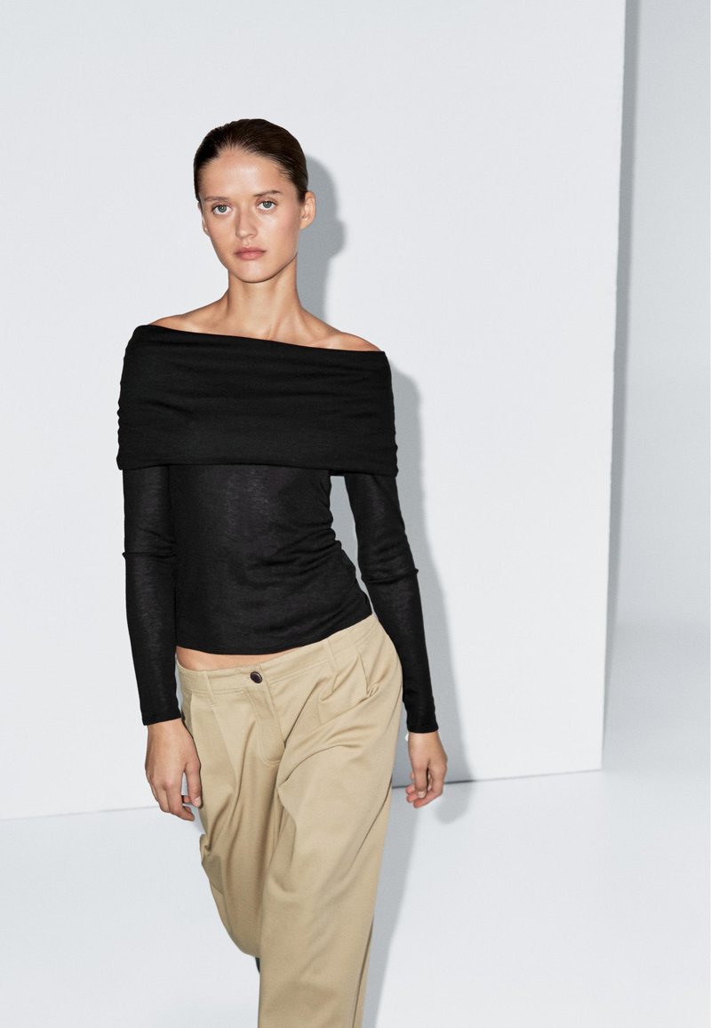 Schwarzes, schulterfreies Langarmoberteil aus einem weichen, leichten Stoff, kombiniert mit beigen, plissierten Hosen mit sichtbarem Knopfdetail.