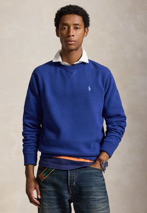 Homme portant un sweat-shirt bleu par-dessus une chemise blanche à col et un jean, avec une main dans la poche et une montre au poignet.