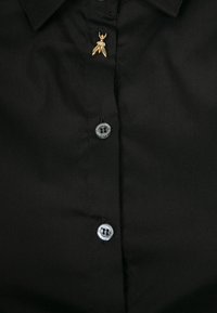 Chemise noire avec deux boutons noirs visibles et une petite broche dorée en forme d'insecte près du col.