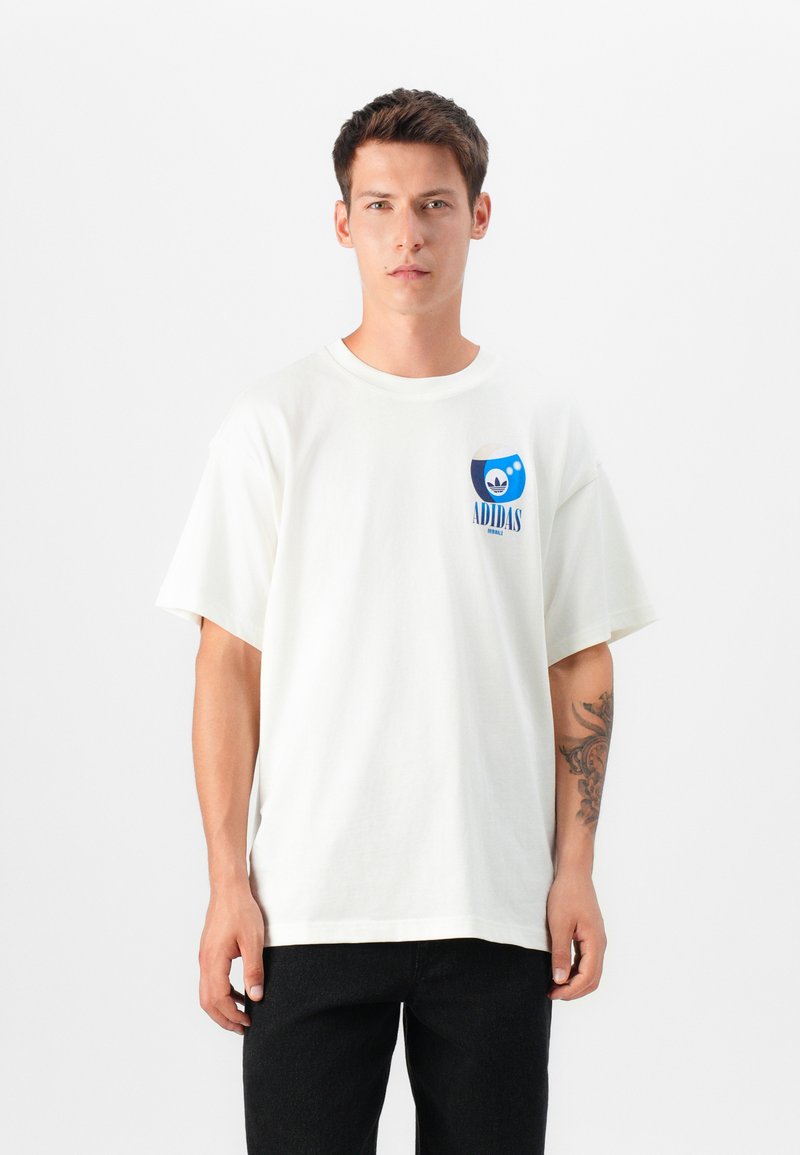 Hvid bomulds t-shirt med korte ærmer, rundudskæring, med et blåt grafisk logo på brystet. Bæret med sorte jeans.