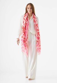 Écharpe en soie rose et orange à motifs drapée sur un chemisier de couleur claire, accompagnée d'un pantalon blanc ample. Le tissu a une texture douce et fluide.