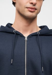 Φούτερ με φερμουάρ σε ναυτικό μπλε χρώμα, από βαμβακερό μείγμα, με μπροστινό φερμουάρ, κορδόνια ρυθμιστικά και απαλό υφή. Χωρίς σχέδια ή πρόσθετες διακοσμήσεις.