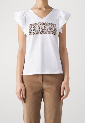 Kobieta ubrana w białą koszulę z dekoltem w serek, z falbankami na rękawach i logo w panterkę, zestawioną z brązowymi, wysokim stanem spodniami, stoi na jednolitym tle.