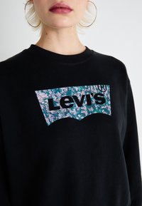 Čierna mikina s veľkým logom Levi’s s kvetinovým modrým a ružovým vzorom. S okrúhlym výstrihom a jemnou textúrou, navrhnutá na každodenné nosenie.